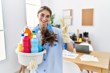 Sticker - Young blonde woman wearing cleaner uniform holding cleaning products smiling with happy face winking at the camera doing victory sign with fingers. number two.