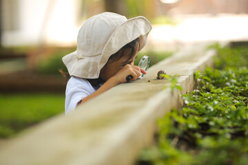 little kid girl asian wearing a white hat and jeans jumpsuit and xploring nature with a magnifying g