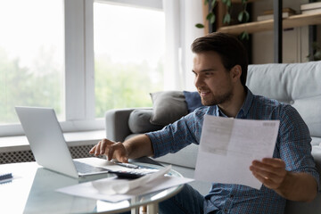 Smiling focused millennial man satisfied with income, savings calculating taxes, budget, costs, paying bill, insurance, mortgage fee, checking paper invoice, doing accounting work on laptop at home