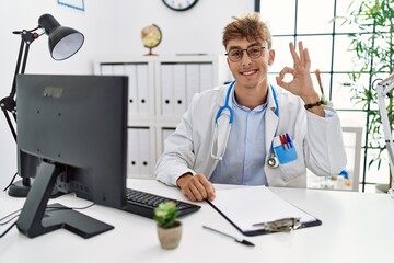 Sticker - Young caucasian doctor man working at the clinic smiling positive doing ok sign with hand and fingers. successful expression.