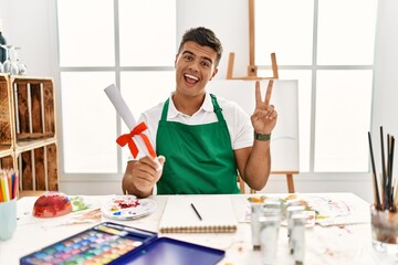 Poster - Young hispanic man at art studio holding degree smiling with happy face winking at the camera doing victory sign with fingers. number two.