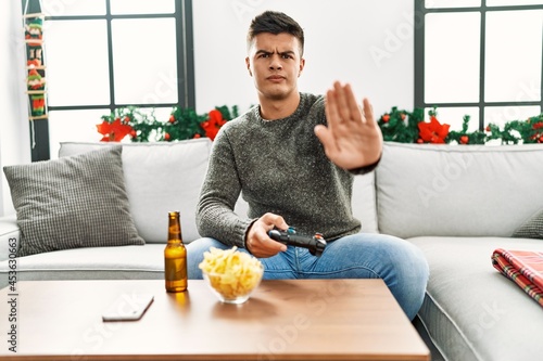 Young hispanic man playing video game on christmas with open hand doing stop sign with serious and confident expression, defense gesture