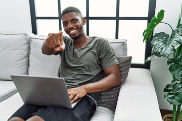 Sticker - Young african american man using laptop at home sitting on the sofa pointing to you and the camera with fingers, smiling positive and cheerful