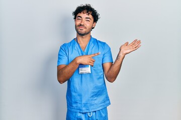 Sticker - Young hispanic man wearing blue male nurse uniform amazed and smiling to the camera while presenting with hand and pointing with finger.