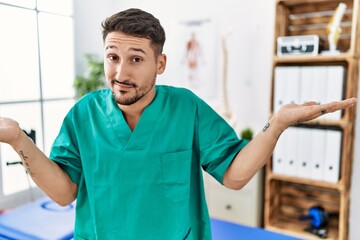 Poster - Young physiotherapist man working at pain recovery clinic clueless and confused expression with arms and hands raised. doubt concept.