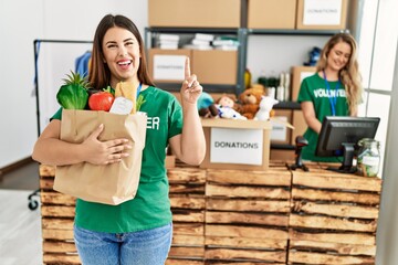 Poster - Young brunette woman at wearing volunteer t shirt holding bag with food surprised with an idea or question pointing finger with happy face, number one