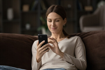 Close up satisfied young woman using smartphone, sitting on couch at home, pleasant young female looking at phone screen, typing writing message in social network, browsing mobile device apps