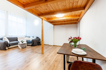 Wall Mural - Bright loft apartment with large window, wooden floor and built-in wooden second floor above. Room has decorative coffee table, big corner sofa with pillows, wall mounted light and set dining table.