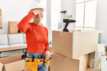 Canvas Print - Middle age grey-haired woman wearing hardhat standing at new home smiling and laughing with hand on face covering eyes for surprise. blind concept.