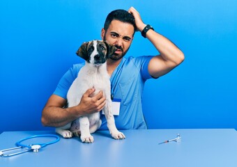 Wall Mural - Handsome hispanic veterinary man with beard checking dog health confuse and wondering about question. uncertain with doubt, thinking with hand on head. pensive concept.