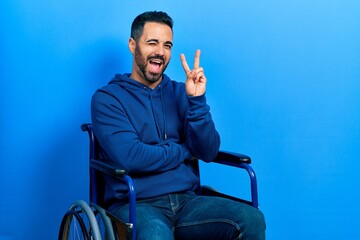 Sticker - Handsome hispanic man with beard sitting on wheelchair smiling with happy face winking at the camera doing victory sign. number two.