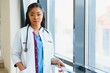 © Serhii - Portrait confident African American female doctor medical professional writing patient notes isolated on hospital clinic hallway windows background. Positive face expression