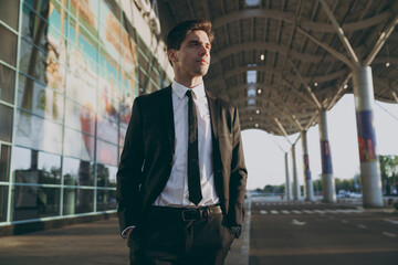 Wall Mural - Bottom view minded young traveler brunet businessman man 20s in black classic tie suit standing outside at international airport terminal look aside. People air flight business trip lifestyle concept.