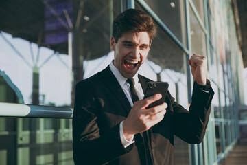 Wall Mural - Young traveler businessman man in black suit stand outside at international airport terminal use mobile cell phone book taxi order hotel do winner gesture clench fist Air flight business trip concept.