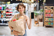 © ViDi Studio - Young woman in casual clothes shopping at supermaket store with craft paper package with groceries show mobile cell phone with blank screen workspace area inside hypermarket. Gastronomy food concept