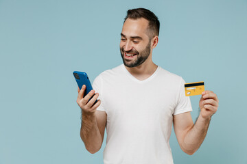 Wall Mural - Young man in white blank print design t-shirt using mobile cell phone hold credit bank card shop online order delivery book tour isolated on plain pastel light blue color background studio portrait