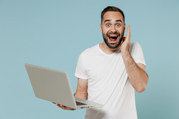 Wall Mural - Young shocked surprised excited happy fun man 20s wearing casual basic white t-shirt hold use work on laptop pc computer hold face isolated on plain pastel light blue color background studio portrait