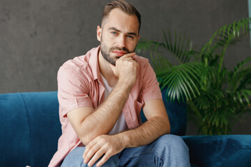 Poster - Young pensive minded confident thoughtful shrewd man 20s in casual clothes looking camera sitting on blue sofa at home flat indoors rest relax on weekends free time. People lounge lifestyle concept