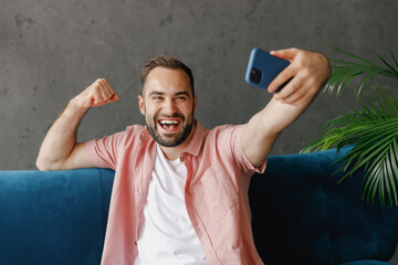 Wall Mural - Young happy man in casual clothes do selfie shot on mobile cell phone post photo social network talk video call do winner gesture sit on blue sofa at home flat indoor rest relax on weekends free time