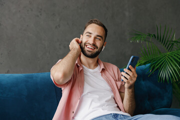 Wall Mural - Young smiling happy calm man 20s wear casual clothes earphones listen to music hold in hand use mobile cell phone look aside sitting on blue sofa at home flat indoors rest relax on weekends free time