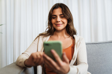 Happy Latin lady relax at home alone sit on room pose share good news at social media via cellphone. Smiling woman enjoy weekend order goods food online in phone app.