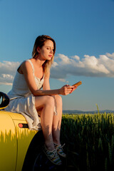 Wall Mural - young girl in sunglasses with smartphone is sitting on a car on green wheat field in spring time