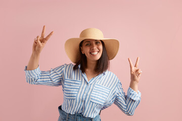 Smiling positive woman with dark hair shows peace sign with both hands, feels excited to go on vacation, wears straw hat, striped shirt, jeans with belt, on pink background. Summer emotions concept.