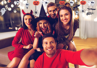 Poster - friendship, holiday and people concept - group of happy smiling friends in halloween costumes of vampire, devil, witch and cheetah taking selfie at home party at night