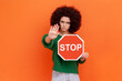 Serious woman with curly hair wearing green casual style sweater showing stop gesture with hand holding red traffic sign of prohibition, blocking. Indoor studio shot isolated on orange background.