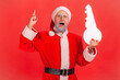 © khosrork - Portrait of amazed elderly man with gray beard wearing santa claus costume standing with open mouth, holding paper key, has idea for buying property. Indoor studio shot isolated on red background.