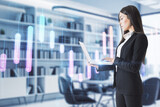 Fototapeta  - Attractive young european businesswoman using laptop in blurry office interior with creative glowing business graph and map. Global investment, trade and profit concept. Double exposure.