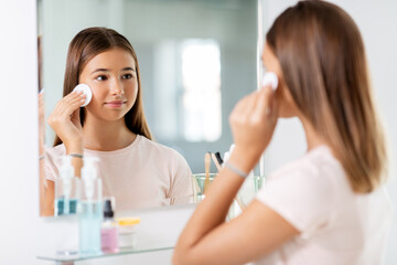 Poster - beauty, hygiene and people concept - teenage girl looking in mirror and cleaning face skin with cotton disc at bathroom