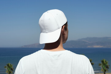Young man in a white baseball cap and white t-shirt on the background of the sea and palm trees back view, baseball cap mock up front view