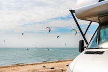Panoramic View Of Many Surf Board Kite Riders On Sand Beach Watersport Spot On Bright Sunny Day Against Rv Camper Van Vehicle At Sea Ocean Coast At Surfing Camp. Fun Adventure Travel Sport Acitivity