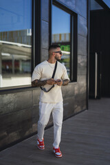 Sticker - Cool caucasian guy with sunglasses and tattoos standing on the background of a building with a phone