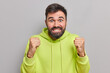 © WHstudio Leushin N - Positive excited bearded man clenches fists roots for favorite football team anticipates results cannot wait for something dressed in sweatshirt poses against grey background. Human emotions