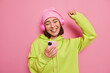 © wayhome.studio  - Joyful upbeat young Asian woman holds modern mobile phone listens favorite music in headphones dances carefree keeps arm raised wears casual green hoodie and hat poses against pink background