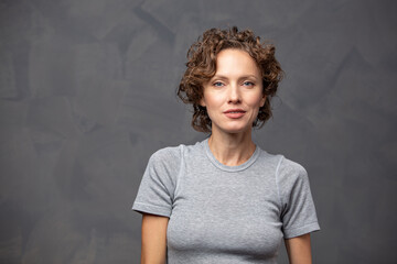 portrait of a serious brunette woman with curly hair wearing casual attire over grey background
