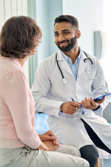 Vertical shot of young happy cheerful Indian medical worker therapist in white doctor's robe having appointment consulting older female patient in modern clinic hospital. Medical healthcare concept.