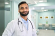 © insta_photos - Portrait of happy friendly male Indian latin doctor medical worker wearing white coat with stethoscope around neck standing in modern private clinic looking at camera. Medical healthcare concept.