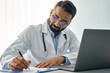 © insta_photos - Indian male medical worker in modern clinic wearing eyeglasses and white doctor's coat using laptop computer writing health personal data, consulting remotely. Telemedicine healthcare concept.