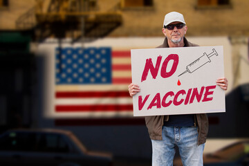 Displeased man with cap, blue jeans and sunglasses holding a NO COVID vaccine sign and a syringe with american stars and stripes flag on a wall in the background. Supporting anti-vaccination movement.