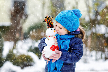 Wall Mural - Cute little toddler girl making mini snowman and eating carrot nose. Adorable healthy happy child playing and having fun with snow, outdoors on cold day. Active leisure with children in winter