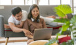 © Bangkok Click Studio - Young attractive Asian couple sitting and looking at laptop together in living room. Woman sitting on the floor wearing headphones and man lying on couch. Concept for love and couple relationship