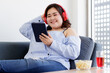 © Bangkok Click Studio - Asian young happy pretty friendly fat plus size woman sit smiling relaxing on gray fabric couch listening to music from big red headphones use playlist from black tablet on hand in living room