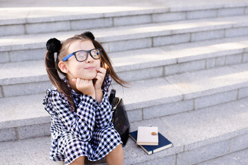 Wall Mural - Funny pretty kid girl wear glasses and stylish dress sitting on stairs with books, backpack outdoors. Cheerful pupil toddler having fun. Back to school. Childhood.