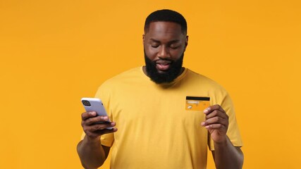 Wall Mural - Cheery vivid young bearded african american man 20s wears orange t-shirt using mobile cell phone hold in hand credit bank card doing online shopping isolated on plain yellow background studio portrait