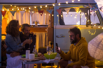 Group of friends enjoy leisure night together with food and drins for dinner outside the van camper vehicle - travel and vacation summer lifestyle with men and woman having fun together