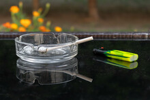 A Transparent Ashtray With A Lit Cigarette Reflected On A Black Glass Table And A Yellow Lighter; Flower Background; Smoking Kills