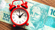 © Rmcarvalhobsb - Real - BRL, dinheiro do Brasil. Um pequeno relógio despertador de cor vermelha sobre cédulas de Real Brasileiro. Conceito de aposentadoria, planejamento financeiro e investimentos.